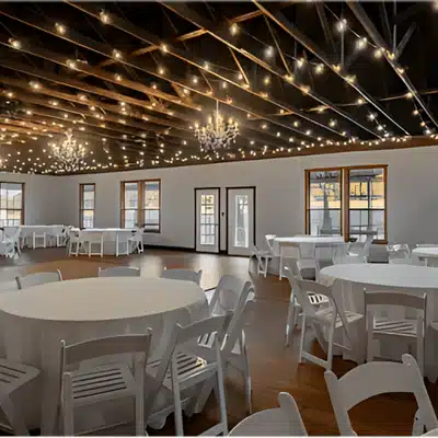 Elegant indoor hall prepared for cocktail hour wedding with round tables, white chairs, and string lights