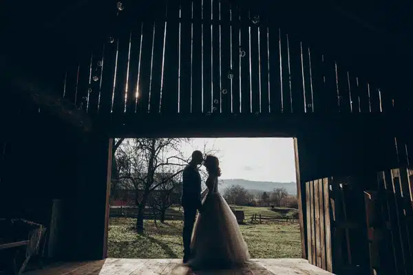 Silhouetted bride and groom kiss inside rustic barn doorway overlooking rural fields at sunset outside