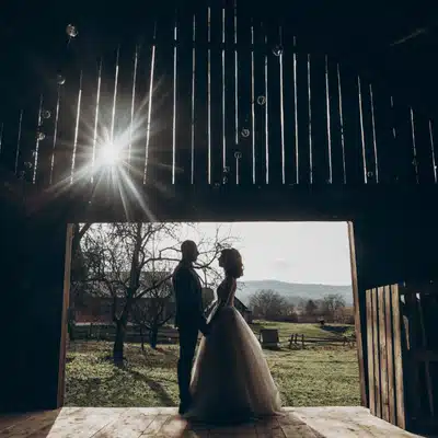 Bride and groom stand hand in hand inside barn doorway at sunset, overlooking peaceful countryside fields.