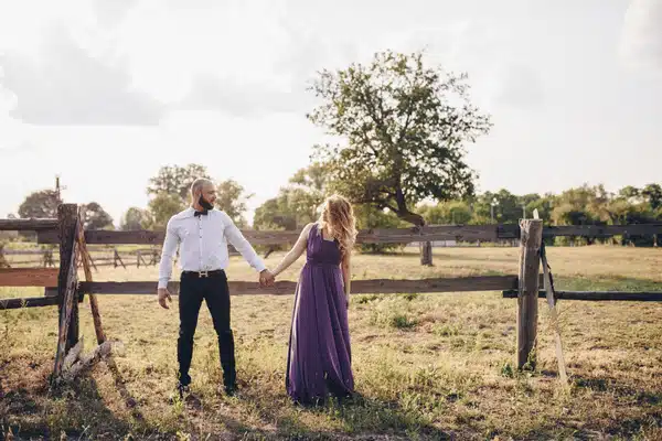 Couple holds hands by rustic wooden fence in open Texas pasture during golden-hour engagement photos