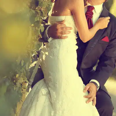 Groom embraces bride in lace gown outdoors, intimate wedding portrait with soft golden light background